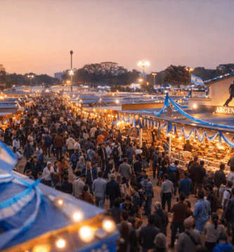 Pasillos llenos y stands iluminados en la Feria del Libro de Kolkata al atardecer; pabellón del país tema con colores celeste y blanco.