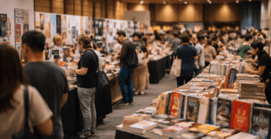 Feria del libro en interior con stands de editoriales y asistentes revisando libros bajo luz cálida.