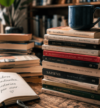 Pila de libros pendientes con una taza y un cuaderno en una biblioteca personal.