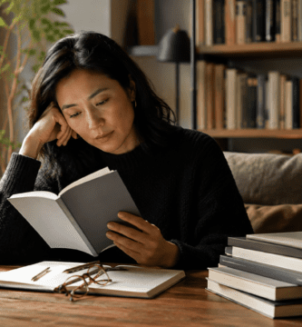 Persona lee una novela corta en una biblioteca cálida con libros apilados sobre la mesa.