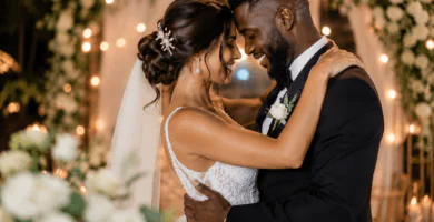 Pareja de novios abrazándose con alegría en una boda romántica con luces cálidas, flores blancas y ambiente elegante.