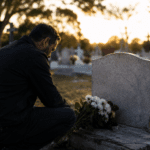 Hombre recordando a su papá fallecido frente a una tumba con flores en un cementerio al atardecer
