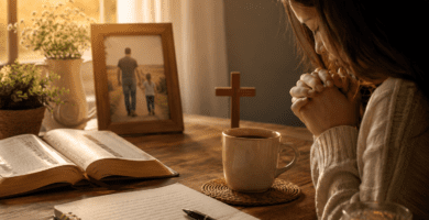 Mujer ora por su papá junto a una Biblia abierta, una cruz y una foto familiar en un ambiente cálido de fe.