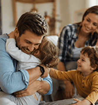 Esposo abrazando a sus hijos en casa mientras su pareja lo mira con amor, imagen cálida sobre un buen padre presente.