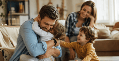 Esposo abrazando a sus hijos en casa mientras su pareja lo mira con amor, imagen cálida sobre un buen padre presente.