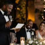 Padrino de boda leyendo un discurso emotivo para los novios durante una recepción elegante con luces cálidas y flores blancas.