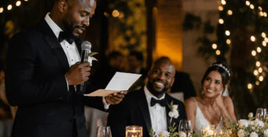 Padrino de boda leyendo un discurso emotivo para los novios durante una recepción elegante con luces cálidas y flores blancas.