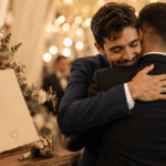 Hermano abrazando al novio en una boda emotiva, junto a una tarjeta y flores en una recepción con luces cálidas.