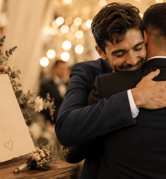 Hermano abrazando al novio en una boda emotiva, junto a una tarjeta y flores en una recepción con luces cálidas.