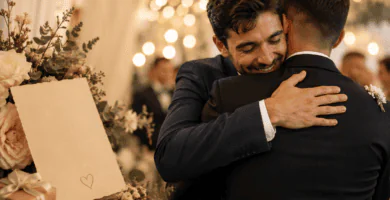 Hermano abrazando al novio en una boda emotiva, junto a una tarjeta y flores en una recepción con luces cálidas.