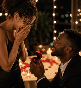 Hombre negro pidiendo matrimonio con un anillo a una mujer emocionada, en una escena romántica con velas, rosas y luces cálidas.