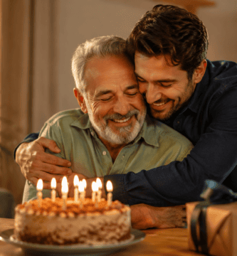 Padre celebrando su cumpleaños con su hijo adulto en casa, entre abrazos, pastel y velas encendidas