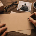 Manos escribiendo una tarjeta para papá junto a un regalo, una fotografía familiar y una taza de café