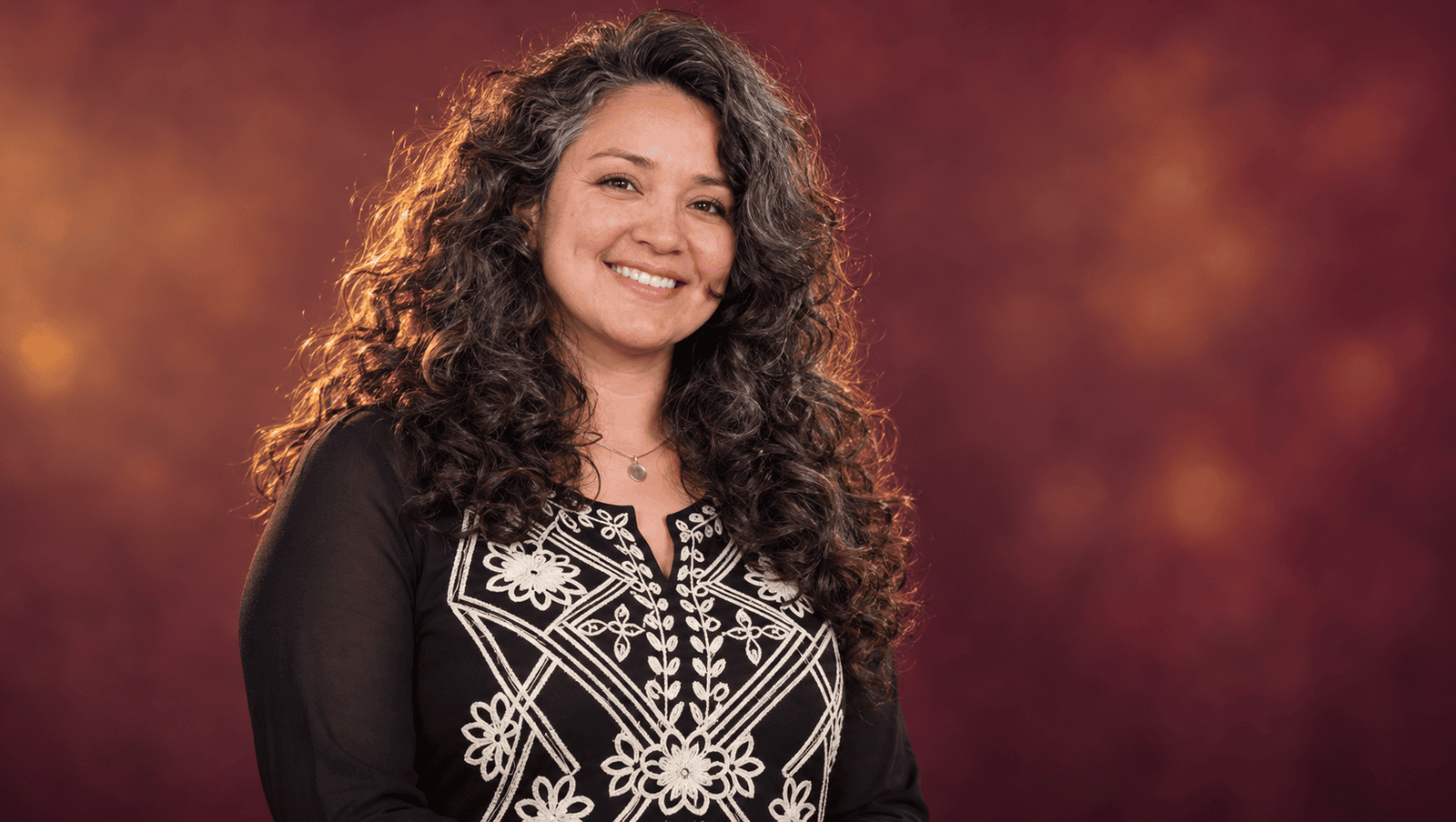 Diana Paola Agámez con cabello rizado y blusa negra bordada sobre un fondo burdeos.