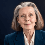 Anne Carson sin lentes, con chaqueta azul petróleo y blusa marfil sobre fondo oscuro.