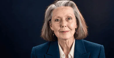 Anne Carson sin lentes, con chaqueta azul petróleo y blusa marfil sobre fondo oscuro.