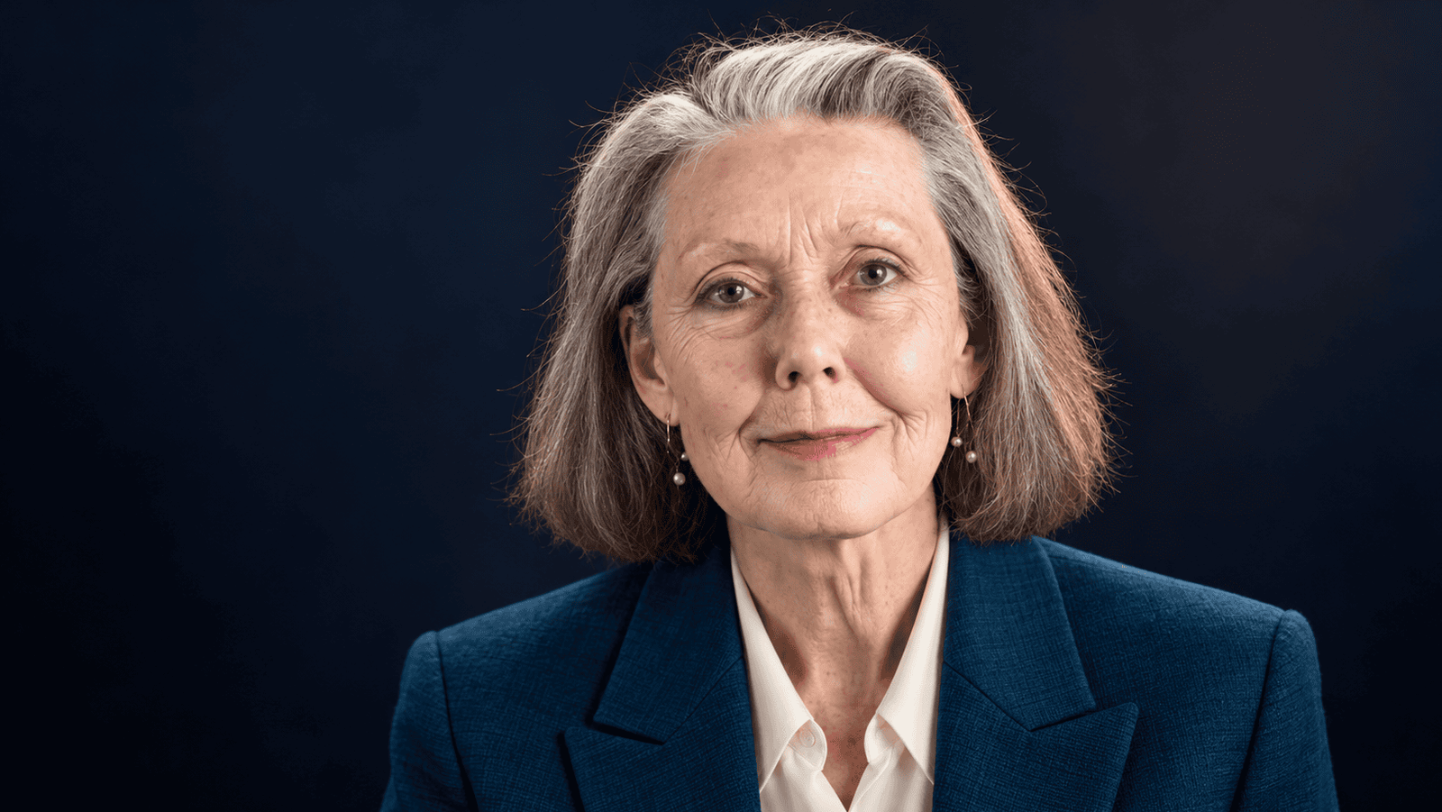 Anne Carson sin lentes, con chaqueta azul petróleo y blusa marfil sobre fondo oscuro.